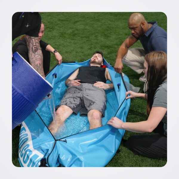 Responders pouring iced water into a blue Polar Life Pod around a man lying on artificial turf